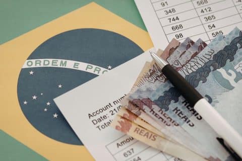 Cédulas de real, caneta e documentos sobre a bandeira do Brasil ilustram pagamentos e valores do INSS